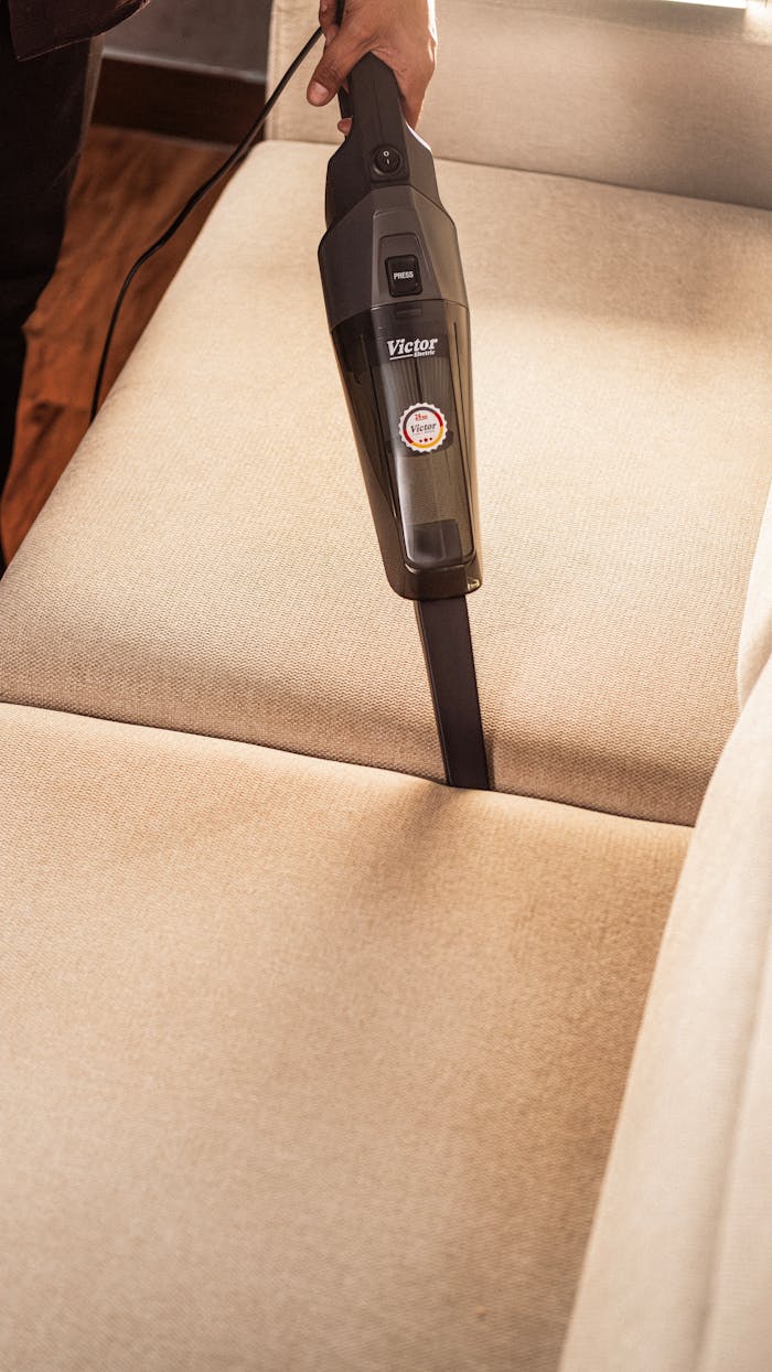 A person using a handheld vacuum to clean a beige sofa indoors, showcasing effective cleaning.